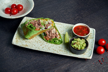Close up view on burrito with beef, rice, tomatoes, corn and bell pepper in green pita on a brown plate with tomato salsa and guacamole. Vegetarian Shawarma Roll