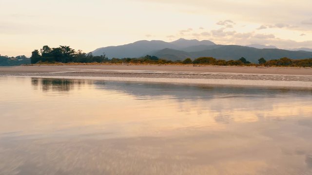 Aerial low above water surface mirroing the colors of sunset and Takaka Hill in the background in Abel Tasman National Park