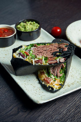 Close up view on burrito with beef, rice, tomatoes, corn and bell pepper in black pita on a brown plate with tomato salsa and guacamole. Vegetarian Shawarma Roll