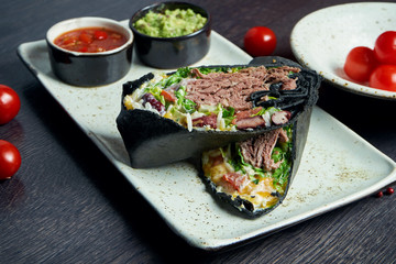 Close up view on burrito with beef, rice, tomatoes, corn and bell pepper in black pita on a brown plate with tomato salsa and guacamole. Vegetarian Shawarma Roll