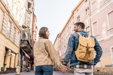 Back view of couple looking at each other and holding hands with backpack in city