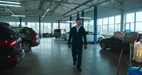 Caucasian young handsome man cars repairment worker in uniform, goggles and with wrench coming close to camera in big garage. Portrait of cool guy mechanic smiling cheerfully and posing.
