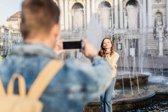 Selective focus of man taking picture with smartphone of woman blowing kiss near fountain in city