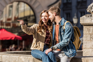 Girlfriend pointing with finger and looking away with boyfriend sitting together on stone and smiling in city