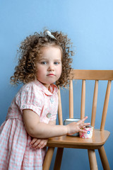 Little curly-haired girl in a pink dress drinks milk from a blue cup of a mug and eats a cupcake
