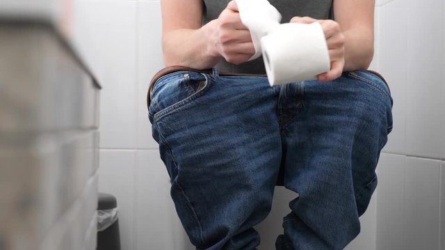 A man with trousers down sitting on the toilet has problems with defecation, he is uptight, he unreels and tear a toilet paper. Slow motion shot.