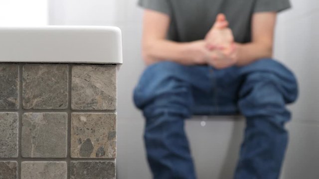 A man with trousers down sitting on the toilet has problems with defecation or kidney stones, he is uptight, clenches his fists and tears trousers.