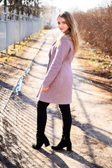 Full-length portrait of a pretty smiling posing blonde Caucasian girl with long beautiful hair outdoors in a pink coat in a park against a tree in the afternoon on a sunny day.