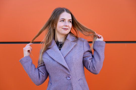 Photo of a cute happy smiling Caucasian blonde girl posing with long beautiful hair outdoors in a pink coat on an orange background in the afternoon on a sunny day.