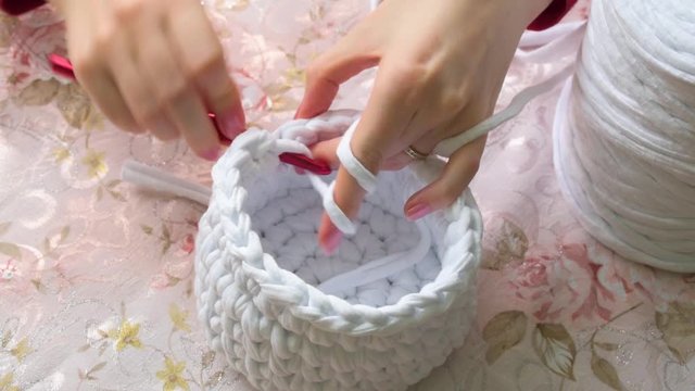 Young woman while crocheting, close up. Stay at home leisure activity idea. White basket made of white T-shirt yarn, with red crochet needle. Pale pink nails. Elegant tablecloth with floral pattern.