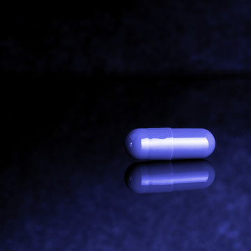 The pill lies on a reflective surface, monochrome image in blue