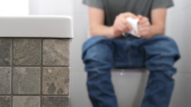 A man with trousers down sitting on the toilet has problems with defecation, he is in pain, holds and crushes a roll of toilet paper, then tear a piece of paper with the intension of using it.