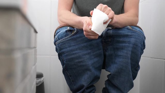 A man with trousers down sitting on the toilet has problems with defecation, he is uptight, holds and crushes a roll of toilet paper, then unreels it and tears.