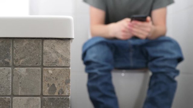 A man with trousers down sitting on the toilet and using a smartphone.