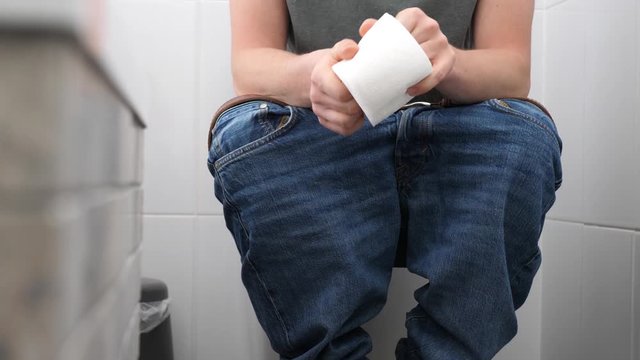 A man with trousers down sitting on the toilet has problems with defecation, he is uptight, holds and crushes a roll of toilet paper, then unreels it and tears. Slow motion shot.