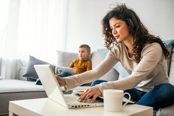 Smiling mom working at home with her child on the sofa while writing an email. Young woman working from home, while in quarantine isolation during the Covid-19 health crisis