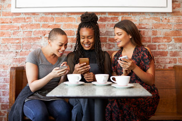 Three Female Friends Meeting For Coffee Sitting At Table Looking At Mobile Phones