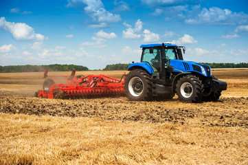 blue new tractor pulls a red harrow raises dust at the field, soil preparation