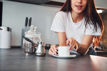 With cup of fresh coffee. Young beautiful brunette in casual clothes indoors in kitchen at daytime