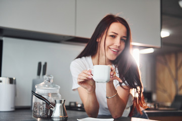 With cup of fresh coffee. Young beautiful brunette in casual clothes indoors in kitchen at daytime
