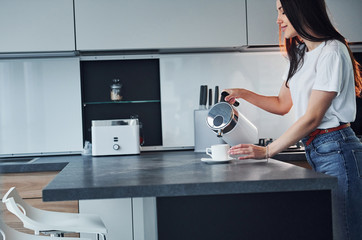 Pouring fresh milk into the glass. Young beautiful brunette in casual clothes indoors in kitchen at daytime