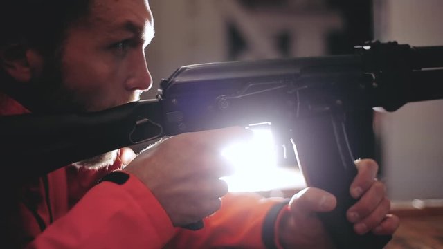 A Caucasian millennial man pulls the trigger of a machine gun at a shooting range at night, a man holds a weapon in his hands, aims, shoots, says, relies on a box, lights in the background.
