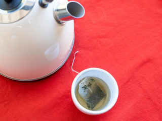 Electric kettle with boiling water for the herbal tea. Tea bag in a white tea cup on a red table,...