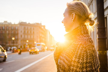 An adult woman in the rays of the setting sun stands on the street and looks away.