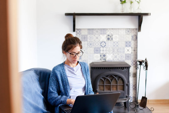 Remote working from home office. Young woman using laptop. Freelancer workplace in living room by wood burning stove, heating. Female business, shopping online, distance education. Lifestyle moment.