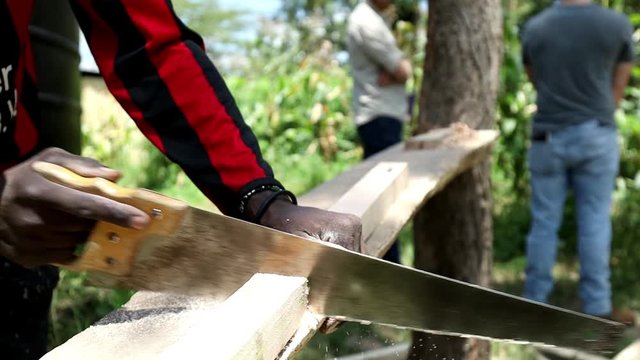 Carpenter using crosscut saw to cut a board to size. Furniture builder in Africa cuts wood to needed specifications to make a new piece for customer. Manual labor, carpentry work in Kenya.