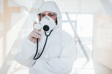 Female doctor scientist in lab coat, defensive eyewear and mask standing indoors and holds stethoscope