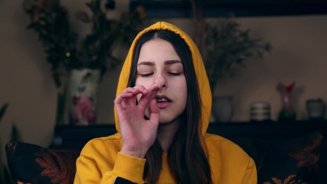 A young girl of European appearance in a brown suit sits at home in an armchair and picks a finger in her nose.