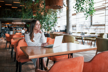 Successful young woman in white dress sits by table with laptop