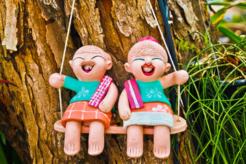 Two smiling ceramic figures of a man and a woman are sitting on a swing, Thailand