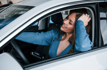 Positive woman in blue shirt sits inside of new brand new car. In auto salon or airport
