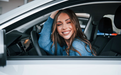 Positive woman in blue shirt sits inside of new brand new car. In auto salon or airport