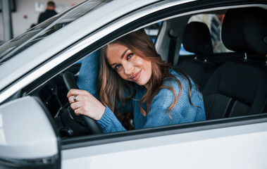 Positive woman in blue shirt sits inside of new brand new car. In auto salon or airport