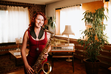 Young beautiful woman smile while playing tenor saxophone in nice cozy condo flat in the sundown...