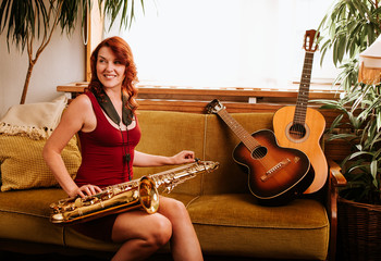 Young beautiful woman sitting on a couch smile while playing tenor saxophone in nice cozy condo...