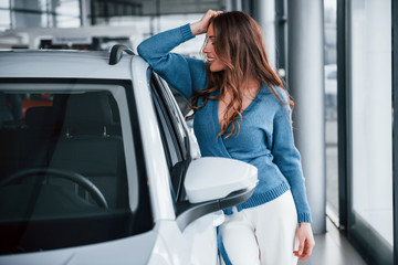 Fototapeta na wymiar Positive woman in blue shirt leaning on brand new car. In auto salon