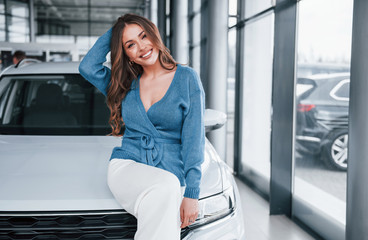 Positive woman in blue shirt sits on brand new car. In auto salon
