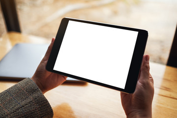 Mockup image of a woman holding black tablet pc with blank white screen with laptop on wooden table
