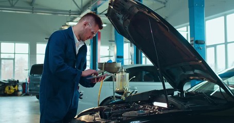 Male Caucasian young auto mechanic standing at open car and repairing motor with tablet device. Man in uniform and goggles working in automobile service with computer as tapping and watching tips.
