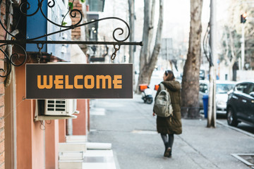 Close-up sign welcome at the entrance to a cafe or shop or another. In the background is a tourist girl with a backpack or a student walking along the street.
