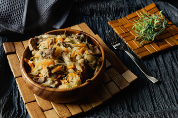 Stewed cabbage with oyster mushrooms and pork tongue with carrots and onions. Hot dish in a wooden plate on a wooden stand, next to a fork, a sprig of grass and a napkin. Black background with relief