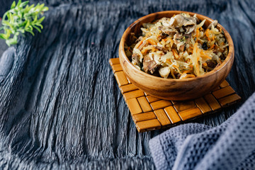 Stewed cabbage with oyster mushrooms and pork tongue with carrots and onions. Hot dish in a wooden plate on a wooden stand, next to a fork, a sprig of grass and a napkin. Black background with relief