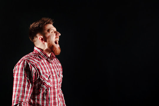 Angry young man in a checked shirt looking to the side sceaming on black background.