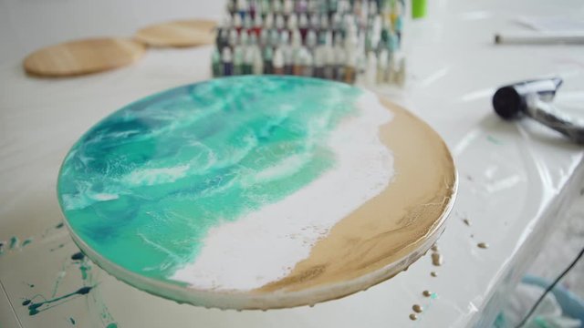 Tracking top view shot of desk with equipments in art studio and unfinished fluid sea painting made with epoxy resin and acrylic paints on round canvas