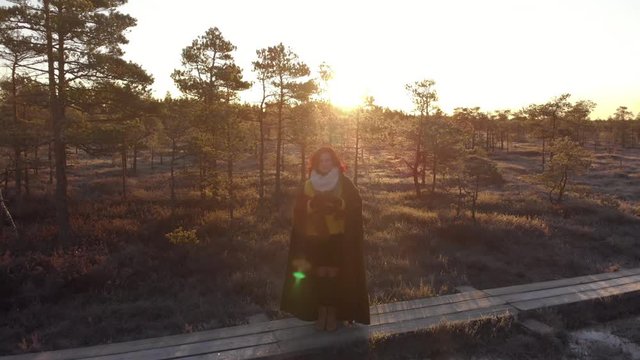 Redhead woman (wears yellow sweater and warm blanket) points big camera at you. Aerial view of beautiful sunrise ar swamp