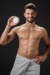 handsome smiling man in towel holding plastic container with face cream isolated on black